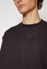 Sweat-shirt noir à col rond en tissu texturé, arborant un logo noir en relief "HUGO" sur la poitrine. Design lisse et minimaliste.