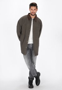 Olivengrøn jakke med en brun ruskinds krave, to brystlommer og trykknaplukninger, parret med grå jeans og sorte støvler.