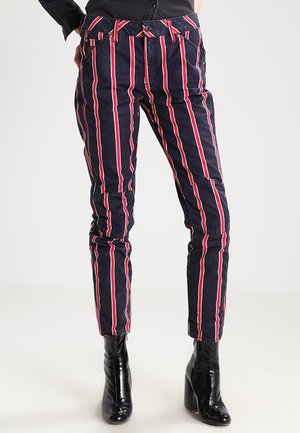 Granatowe spodnie z pionowymi czerwono-białymi paskami, noszone z czarnymi botkami i czarną koszulą wpuszczoną do środka.