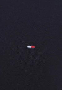 Tejido azul marino con un patrón texturizado. Pequeño logo bordado que presenta tres franjas horizontales: roja, blanca y azul.