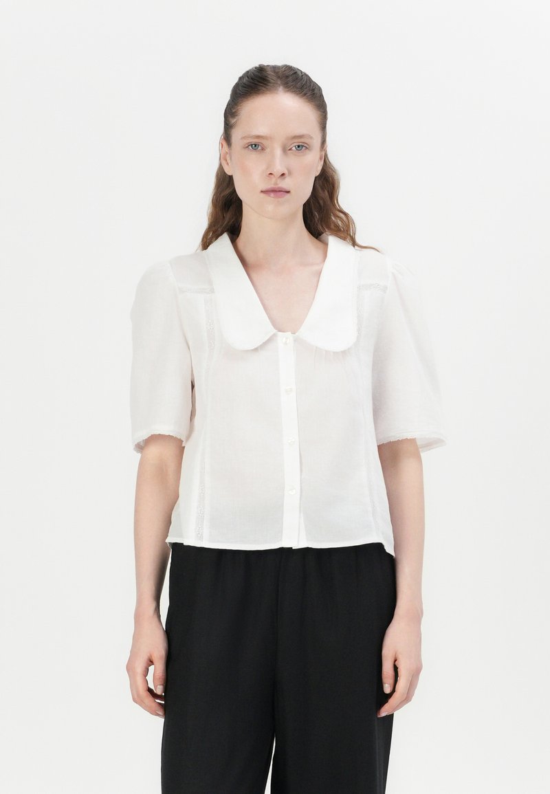 Blusa branca de mangas curtas com um grande colarinho, fecho frontal com botões e textura transparente. Estilizada com calças pretas largas.