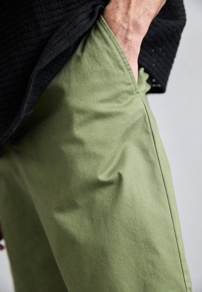 Pantalon en coton vert avec une texture lisse, poches latérales et coupe décontractée, partiellement obscurci par une chemise noire texturée.