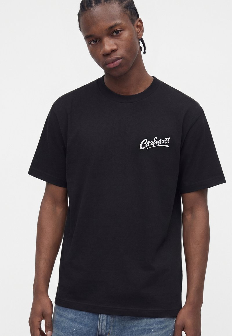 Ung mand med flettet hår iført en sort Carhartt t-shirt og blå jeans, stående mod en ensfarvet lys baggrund.