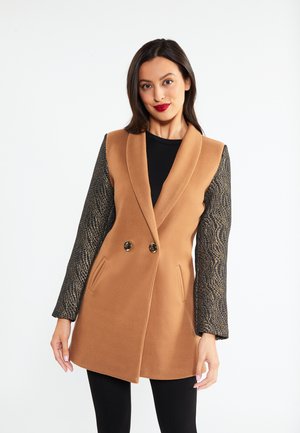 Femeie purtând o vestă camel fără mâneci, cu mâneci texturate negre lungi și două nasturi, pozând pe un fundal alb simplu.