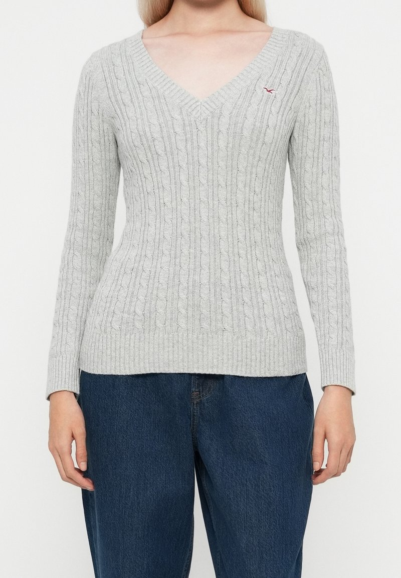 Femme portant un pull en maille torsadée gris clair à col en V et un jean bleu foncé, debout devant un fond blanc uni.