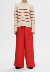 Cremfarbener Pullover mit roten horizontalen Streifen, kombiniert mit lockeren roten Hosen. Der Pullover hat einen runden Ausschnitt und lange Ärmel.