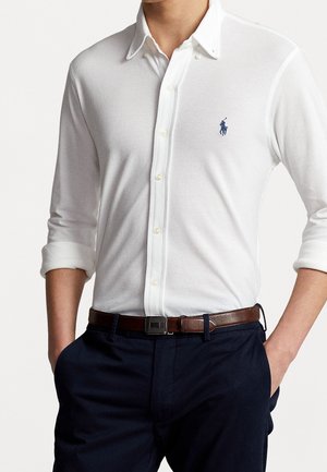 Homme portant une chemise blanche à manches longues avec un logo polo bleu, un pantalon bleu marine et une ceinture en cuir marron, les mains dans les poches.