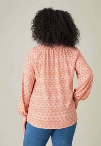 Blusa rossa a fantasia con maniche lunghe a palloncino, scollatura arricciata e tessuto leggero, abbinata a jeans blu in una posa casual.