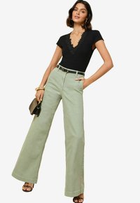 Top nero con bordo in pizzo, pantaloni ampi verde menta a vita alta, cintura nera e sandali con cinturini. Piccola borsa nera tenuta in una mano.