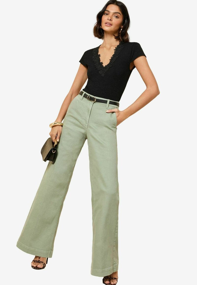 Top nero con bordo in pizzo, pantaloni ampi verde menta a vita alta, cintura nera e sandali con cinturini. Piccola borsa nera tenuta in una mano.