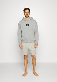 Felpa grigia chiara con un logo nero, caratterizzata da una vestibilità comoda. Abbinata a pantaloni corti grigi chiari, indossati a piedi nudi su uno sfondo semplice.