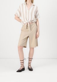Shorts in lino beige abbinati a una camicia a maniche corte bianca e beige a righe, annodata in vita, e ballerine nere con lacci.