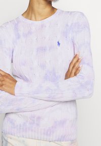 Jasnofioletowy sweter z dzianiny warkoczowej z okrągłym dekoltem, ozdobiony subtelnym wzorem tie-dye i małym niebieskim logo na piersi.