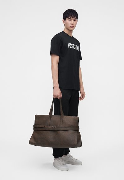 Νέος άνδρας που φοράει μαύρο t-shirt Moschino και μαύρο παντελόνι, κρατώντας μια μεγάλη καφέ δερμάτινη τσάντα, στέκεται μπροστά σε ένα απλό λευκό φόντο.