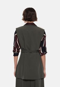 Persona con capelli castani corti che indossa un gilet grigio scuro senza maniche su una camicia a maniche lunghe bordeaux e blu, rivolta di spalle su uno sfondo bianco.