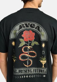 Camiseta negra con un diseño gráfico colorido que presenta una rosa roja, una serpiente, un sol y el texto "PASADO. PRESENTE. FUTURO." en un estilo vintage.