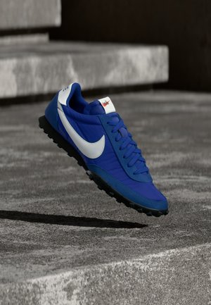 Μπλε αθλητικό παπούτσι Nike με λευκό λογότυπο Swoosh, μαύρη σόλα και κορδόνια, αιωρούμενο πάνω από γκρι σκάλες από σκυρόδεμα σε εξωτερικό χώρο.