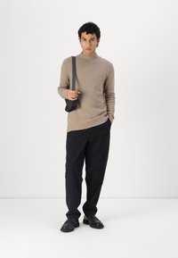 Maglione beige a maglia con un colletto a coste, abbinato a pantaloni neri dalla vestibilità ampia e scarpe nere slip-on; modello che tiene una piccola borsa nera.