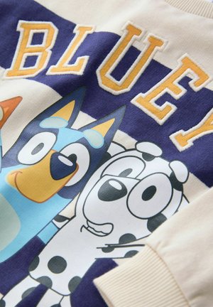 Marškinėliai su mėlyno šuns personažu, turinčiu geltoną nosį, juodai baltu taškuotu šunimi ir žodžiu "BLUEY" geltonomis nėrimo raidėmis ant kreminio fono.