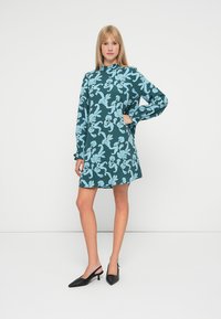 Vestido verde-água com padrão floral em azul claro, mangas longas, gola alta e corte solto. Combinado com sapatos de salto agudo pretos.