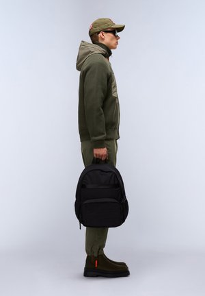 Schwarzer Rucksack aus strukturiertem Stoff mit Fronttasche, gehalten von einer Person in einer olivgrünen Jacke und Hose, die eine Sonnenbrille und eine Kappe trägt.
