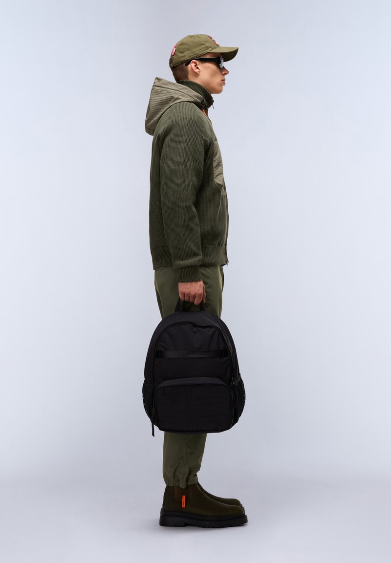 Svart ryggsäck med texturerad tyg och framficka, hålls av en person i en olivgrön jacka och byxor, iförd solglasögon och en keps.