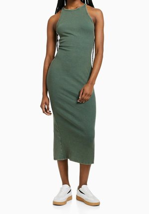 Personne portant une robe midi côtelée sans manches de couleur vert olive, associée à des baskets blanches avec des semelles en gomme, sur un fond blanc.