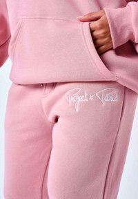 Sudadera de algodón rosa con bolsillo frontal, combinada con pantalones de chándal a juego que presentan el texto bordado en blanco "Project x Paris". Textura suave.
