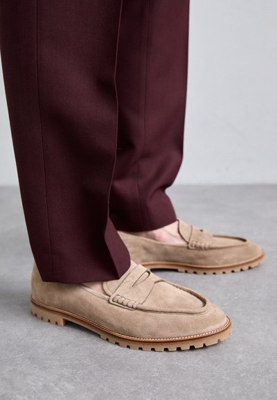 Mocasini bej din suede cu detalii cusute și talpă din cauciuc, asortați cu pantaloni burgundy la comandă, evidențiind textura și contrastul de culoare al pantofilor.