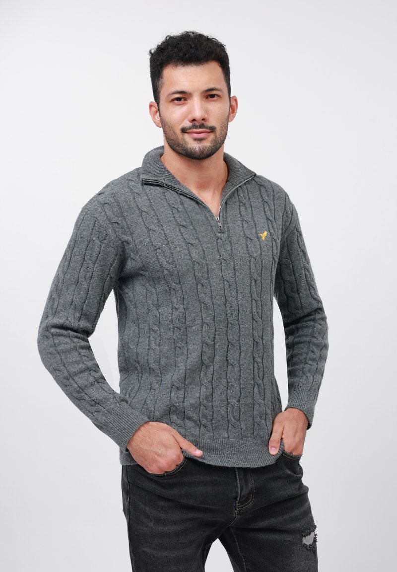 Un uomo sta in piedi con le mani in tasca, indossando un maglione grigio a trecce con zip e jeans scuri strappati, su uno sfondo bianco.