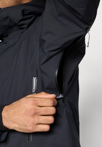 Casaco impermeável azul-marinho com bolso com fecho de correr, capuz ajustável e tecido texturizado. Mão a segurar o puxador do fecho para abrir.