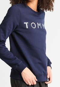 Marinblå crewneck-tröja med vita tryckta bokstäver "TOMMY" som bärs av en person med lockigt hår, med händerna vilande vid höfterna.