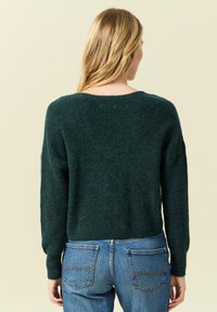 Maglione lavorato a maglia di colore verde scuro con una consistenza morbida e un colletto rotondo, abbinato a jeans blu con tasche posteriori e dettagli di cucitura.