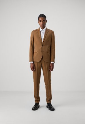 Costume marron ajusté avec une coupe slim, revers crantés et poches latérales, associé à une chemise blanche boutonnée et des chaussures de ville noires.