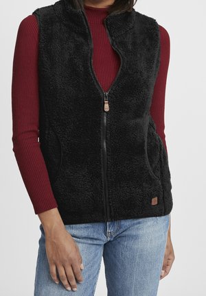 Frau trägt eine schwarze Fleece-Weste mit Reißverschluss über einem langärmligen, gerippten roten Oberteil und hellblauen Jeans, vor einem einfarbigen Hintergrund stehend.