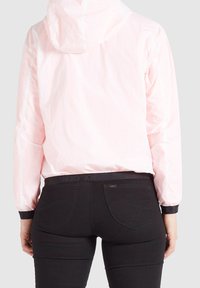 Jasnoróżowa kurtka z kapturem, z elastycznymi mankietami i dołem, noszona na czarnych dżinsach z firmowym paskiem i tylnymi kieszeniami.