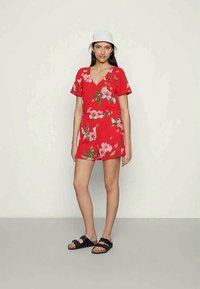 Macacão floral vermelho com mangas curtas e decote em V, apresentando padrões de flores coloridas e uma cintura ajustada, combinado com calçado preto.