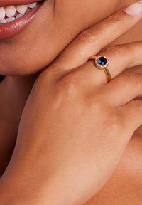 Gouden ring met een ovale blauwe steen omringd door kleine heldere accentstenen, geplaatst op een hand met een licht gepigmenteerde huid.