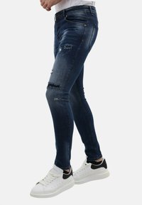 Dunne blauwe denimjeans met slijtage, waaronder scheuren op de knieën en voorzakken. Gecombineerd met witte sneakers met zwarte accenten.