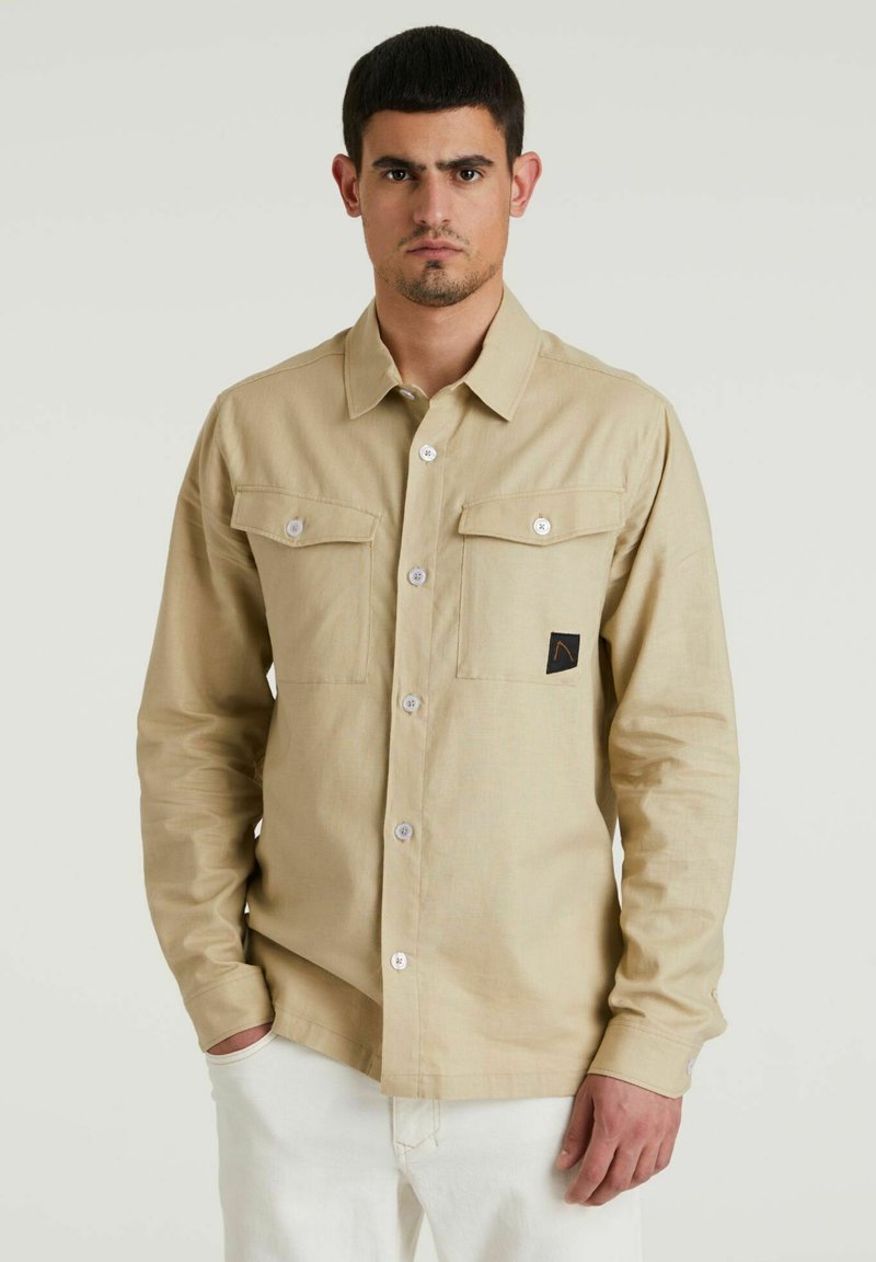 Helles beige Langarm-Hemd mit Knöpfen, zwei Brusttaschen, weißen Knöpfen und einem kleinen schwarzen Logo-Patch. Weicher, strukturierter Stoff.