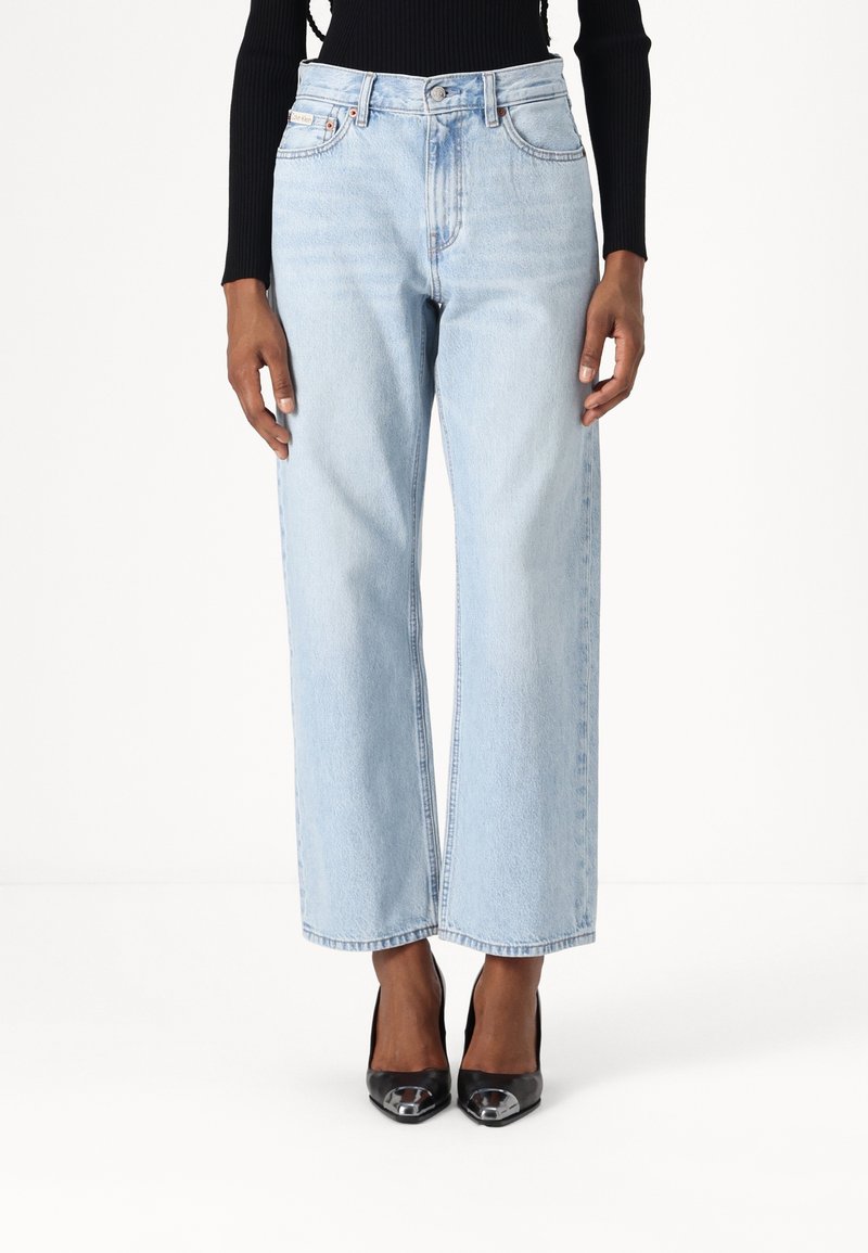 Jeans de mezclilla de tiro alto y pierna ancha en azul claro, con bolsillos frontales y un ligero desvanecimiento. Combinados con tacones negros.