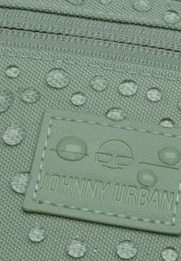 Zelený látkový pouzdro s texturovaným povrchem a kruhovými kapkami vody, s našitým logem a zipovým uzávěrem.