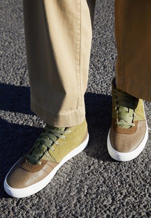 Persona che indossa pantaloni beige e scarpe da ginnastica bicolore verde e marrone in piedi su un terreno asfaltato strutturato.