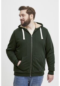 Sudadera con cremallera de color verde oscuro, con un forro beige suave, que presenta cordones blancos, bolsillos frontales y puños de canalé. Tejido casual y texturizado.