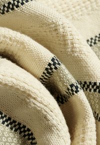 Gros plan sur un tissu tissé de couleur crème avec des motifs texturés et des rayures horizontales noires et grises, montrant des plis et des détails textiles.