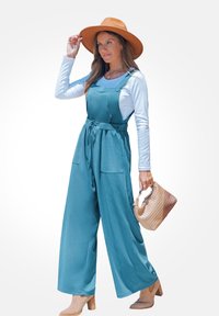 Teal jumpsuit met brede benen, twee voorzakken en knoopdetails. Gecombineerd met een witte blouse met lange mouwen, een tan hoed en een gevlochten tas.