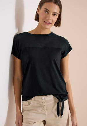 Jeune femme aux cheveux courts et bruns portant un haut noir à manches courtes avec un détail en mesh et un pantalon beige, debout contre un mur clair.