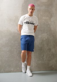 Hvit t-skjorte med svart "ellesse"-logo, blå shorts og hvite sneakers. Glatt tekstur, uformell passform, står mot en betongvegg.