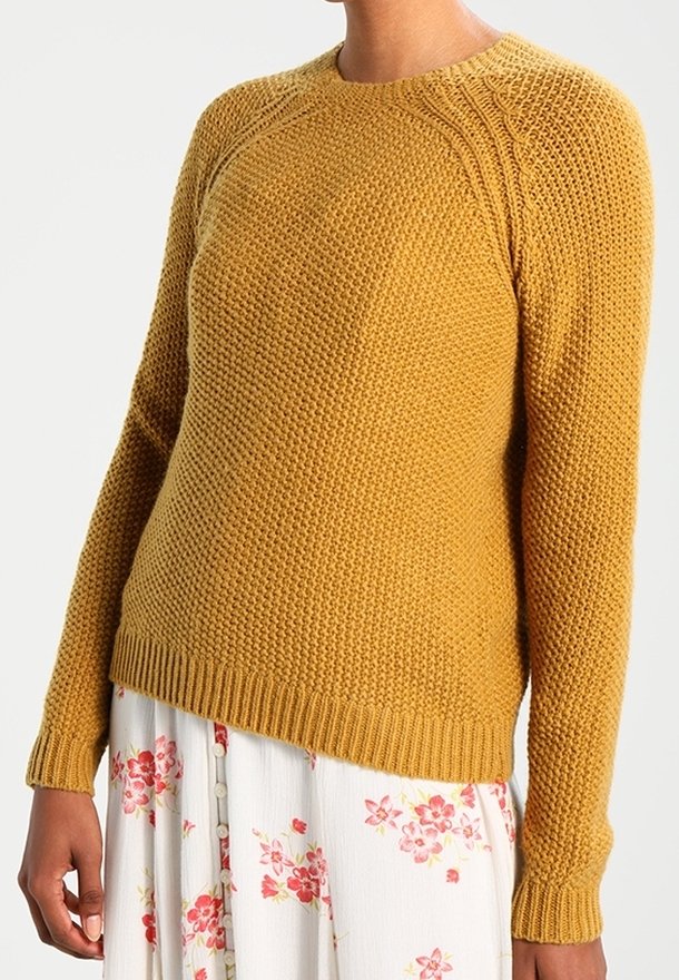 Femme portant un pull en maille texturé jaune moutarde associé à une jupe blanche avec des motifs floraux roses et des boutons.