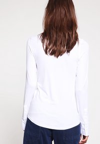 Donna con capelli marroni di lunghezza spalle che indossa una camicia bianca a maniche lunghe e pantaloni blu scuro, vista di spalle su uno sfondo neutro.
